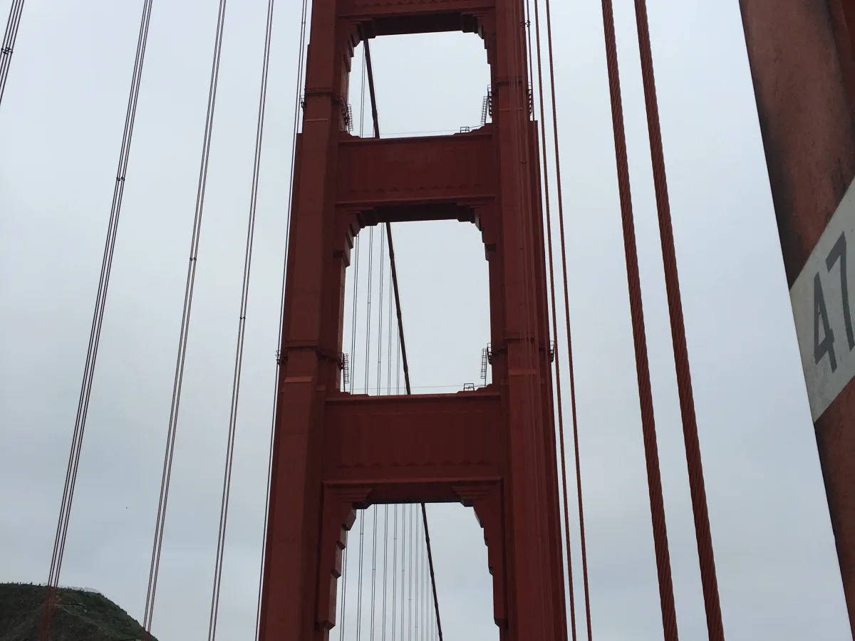 Upward-angled view of a large red suspension bridge tower and vertical cables, with a roadway and pedestrian walkway at the bottom and green hills in the background under an overcast sky.