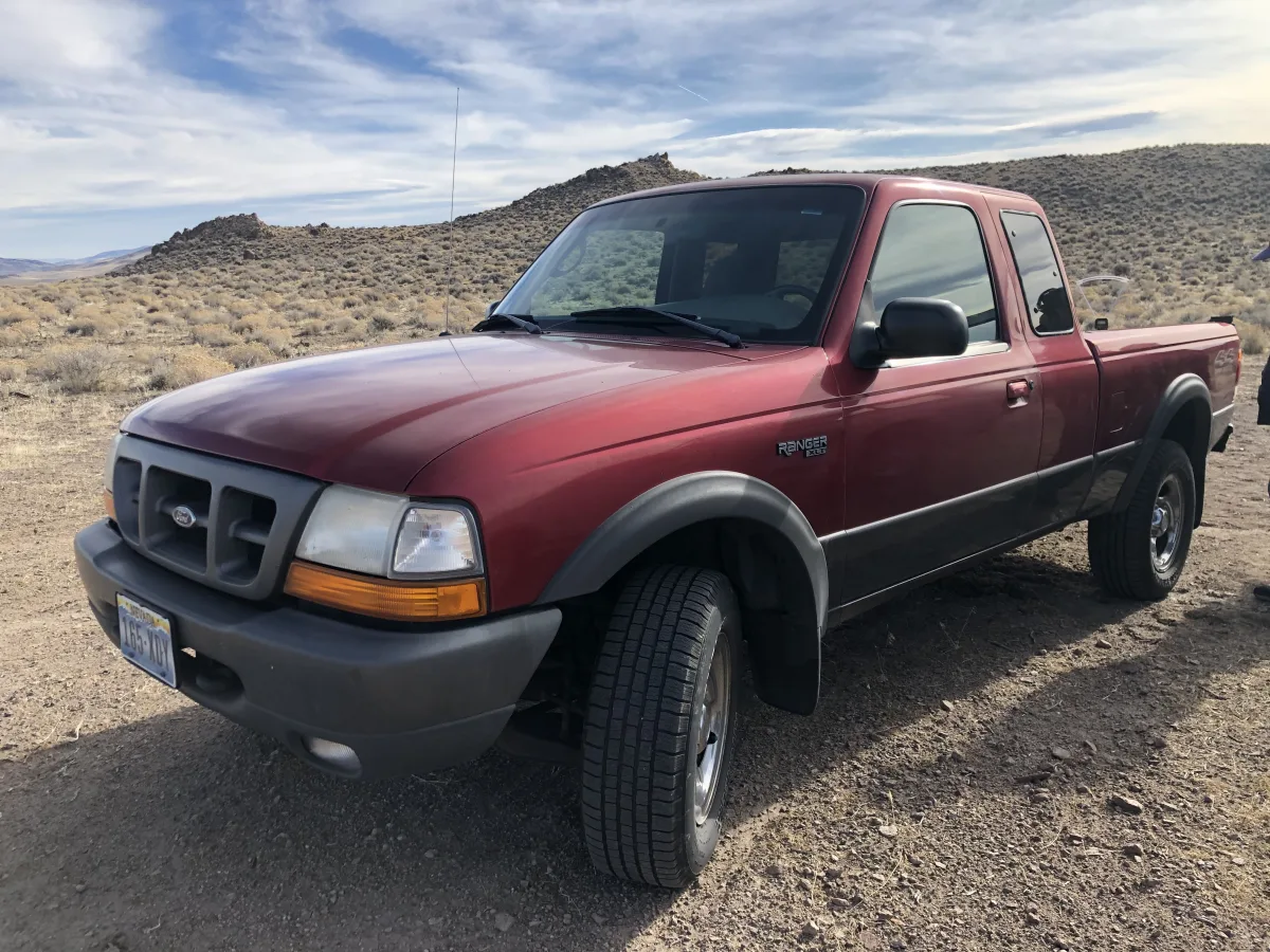 A red pickup truck is parked on a dirt surface in a dry, scrubby landscape with low hills/mountains in the background under a partly cloudy sky.
