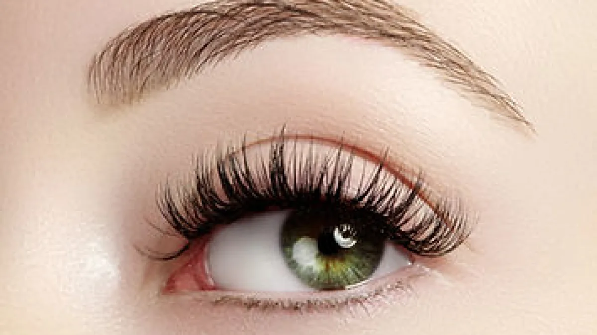 Close-up beauty image focused on a single eye with long eyelashes and a groomed eyebrow; soft neutral background with no visible text.