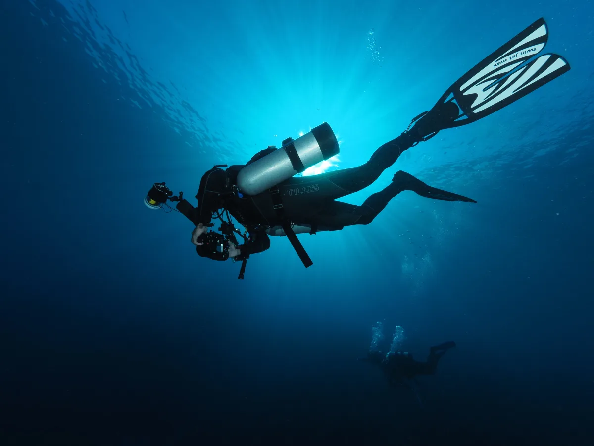 Underwater wide-angle photo showing a scuba diver in a wetsuit swimming horizontally with fins, a single large tank, and an underwater camera rig; sunlight rays stream down through blue water, with another diver faintly visible in the dista