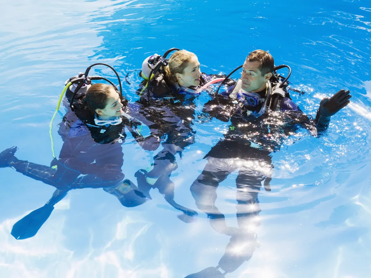 Three people wearing scuba diving gear float at the surface of a bright blue swimming pool, appearing to converse during a training or instruction moment.